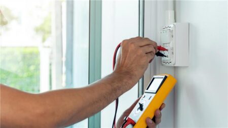 Hand using a multimeter to test wiring in a wall-mounted electrical box near a window with natural light.